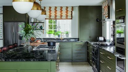kitchen with green cabinets and island, black worksurface, brass globe lights and patterned blinds 