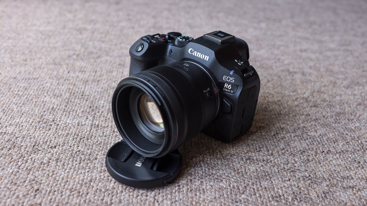 The Canon EOS R6 III with the Canon RF 45mm f/1.2 lens attached and propped up by the lens cover, on a brown carpet.