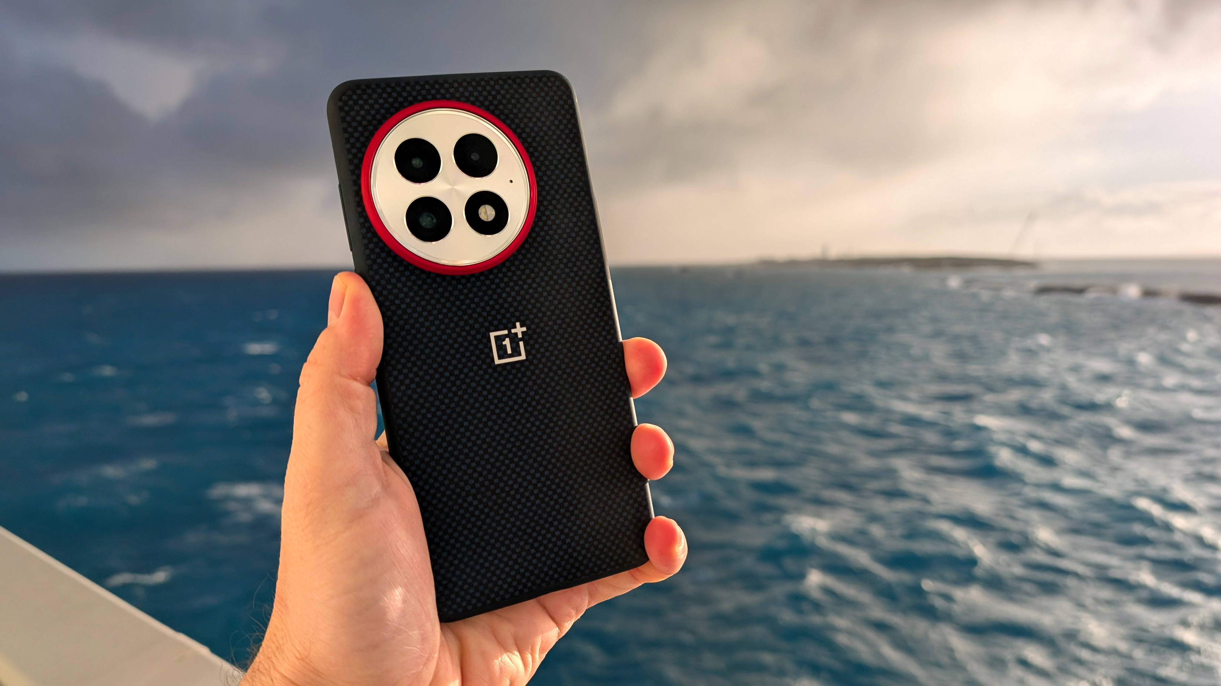 Holding the OnePlus 13 in its blue case with the ocean in the background