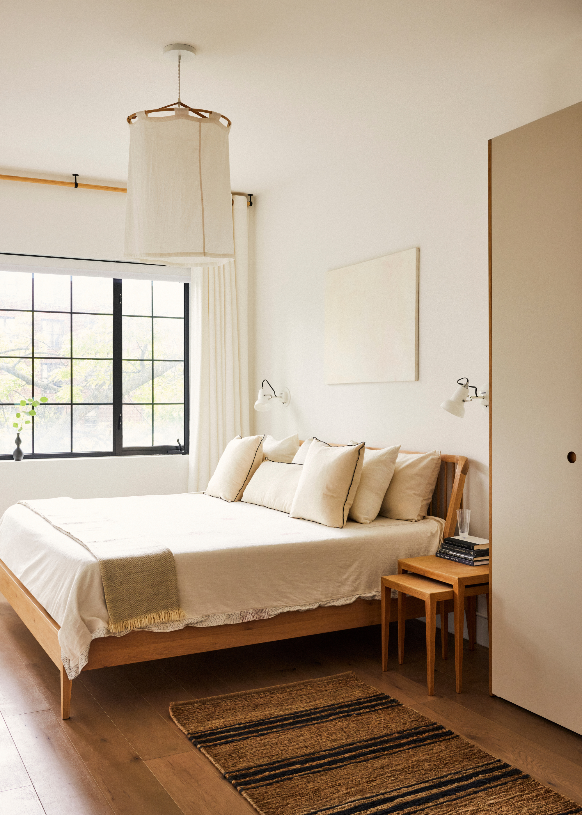 A white bedroom with a wooden bed frame, a white bedscape, a woven runner on the floor, a set of stacked side tables, a pair of white wall sconces, and a hanging lamp shade