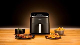 A Cosori air fryer sitting on a wooden work surface surrounded by plates and bowls of food