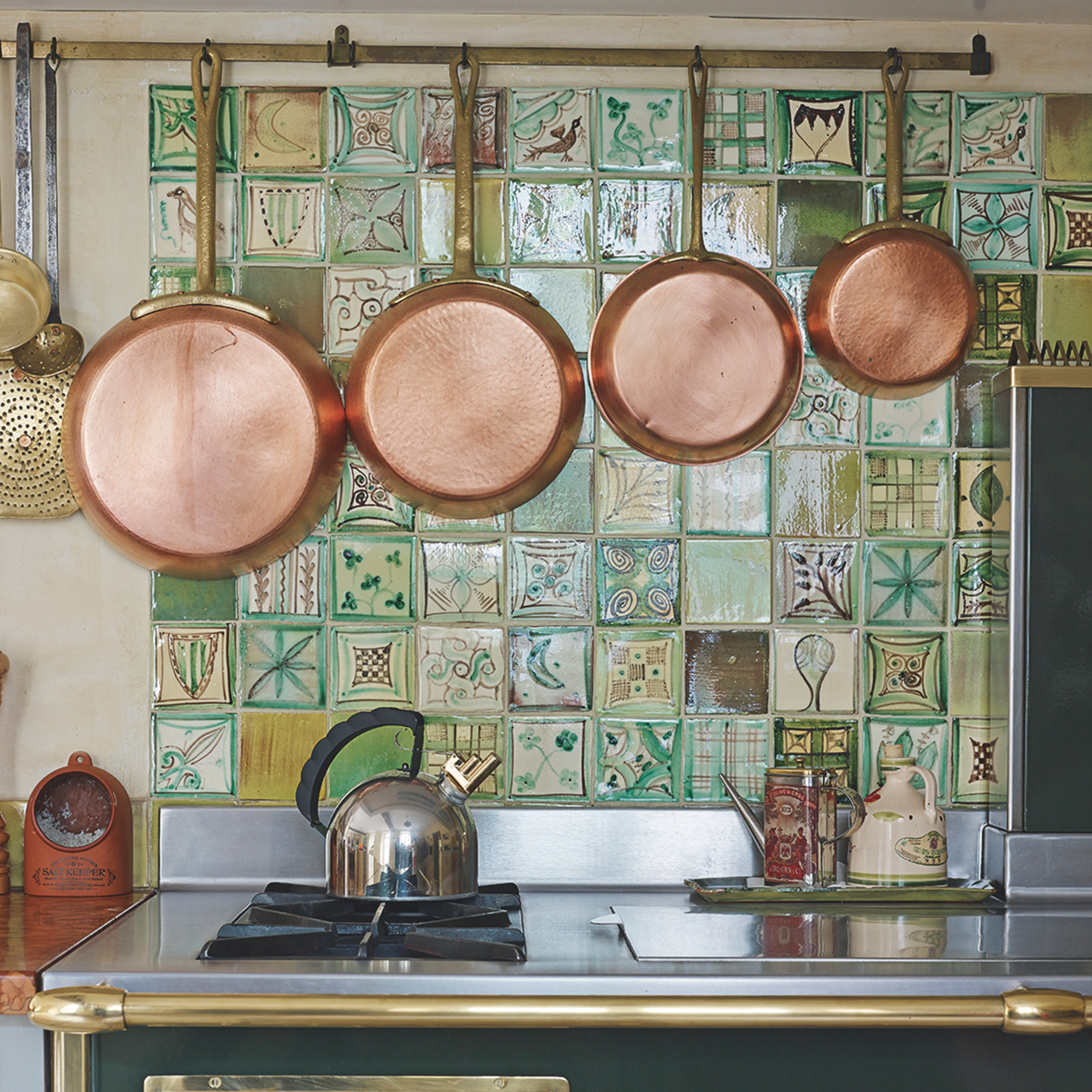 copper pans on rail in front of a patterned green tiled splashback and above a green and gold range cooker