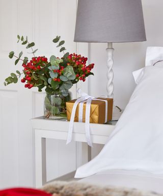 A white bed with a white bedside table, topped with a lamp, a vase of red flowers, and a wrapped gift