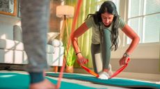 Woman in sportswear kneels down and holds both ends of a long resistance band, her front foot is on the middle of the resistance band. She is in a domestic setting with a couch in the background and someone's lower leg in the foreground