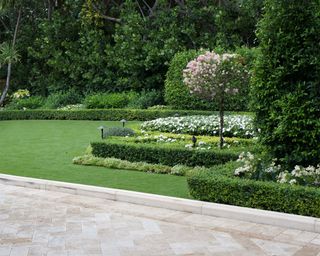 Front yard flower bed ideas surrounded by low hedges in a large, grassy yard lined with trees.
