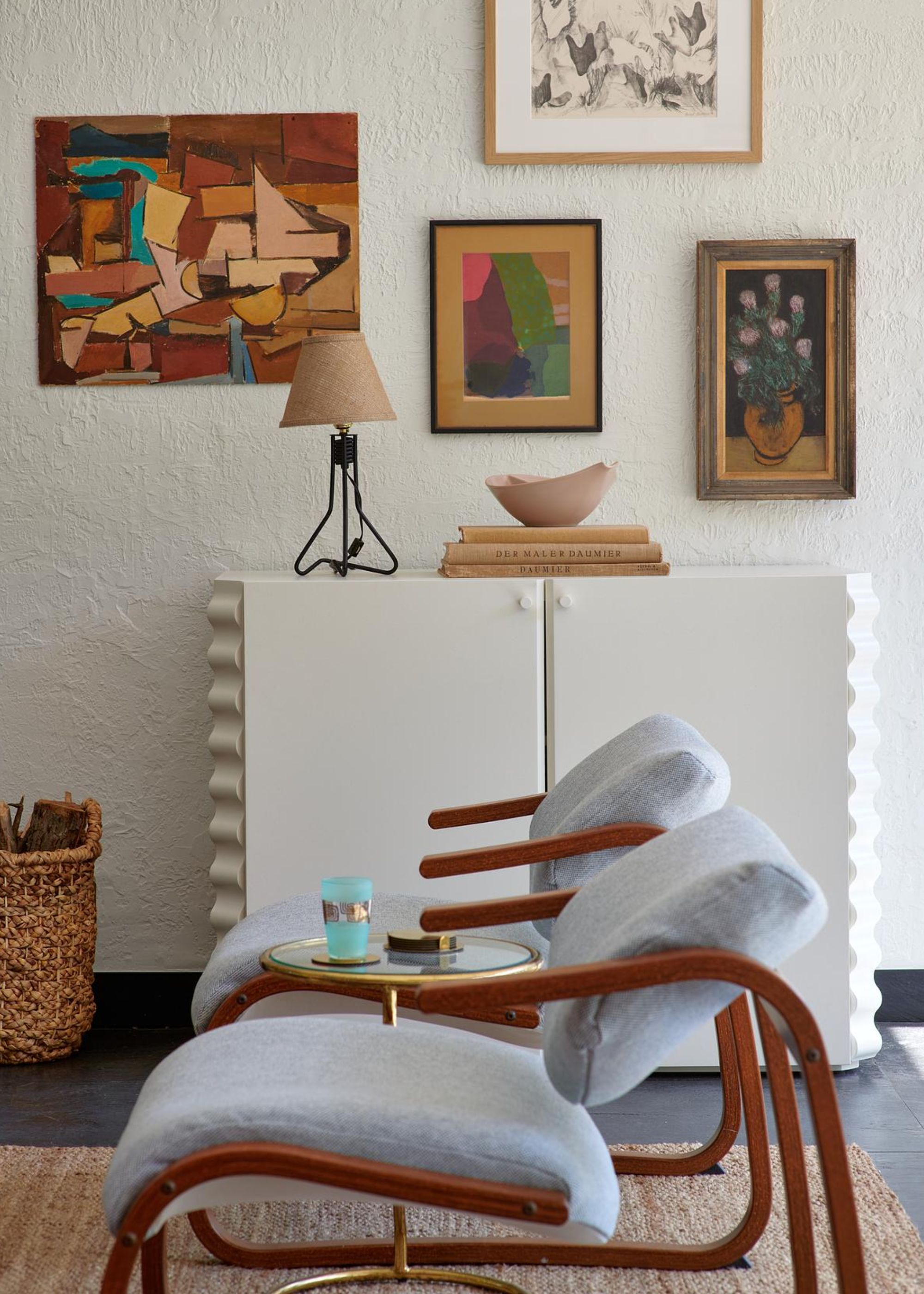 A white living room with colorful art, a textured white cabinet, a table lamp, a woven log basket, and a pair of blue wood armchairs