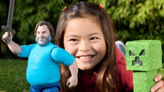 A young girl plays and holds a toy in each hand, one is a Jack Black action figure, the other a green Creeper from Minecraft