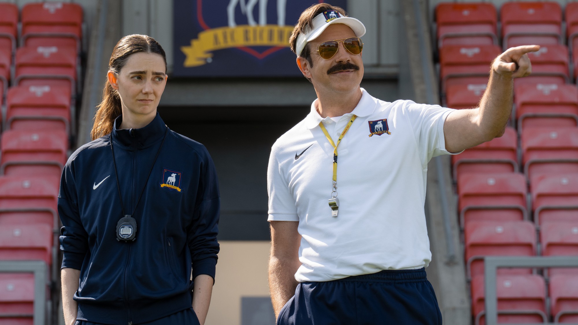 From left to right: Tanya Reynolds standing with her hands in her pockets and Jason Sudeikis pointing with his right hand in Ted Lasso. 