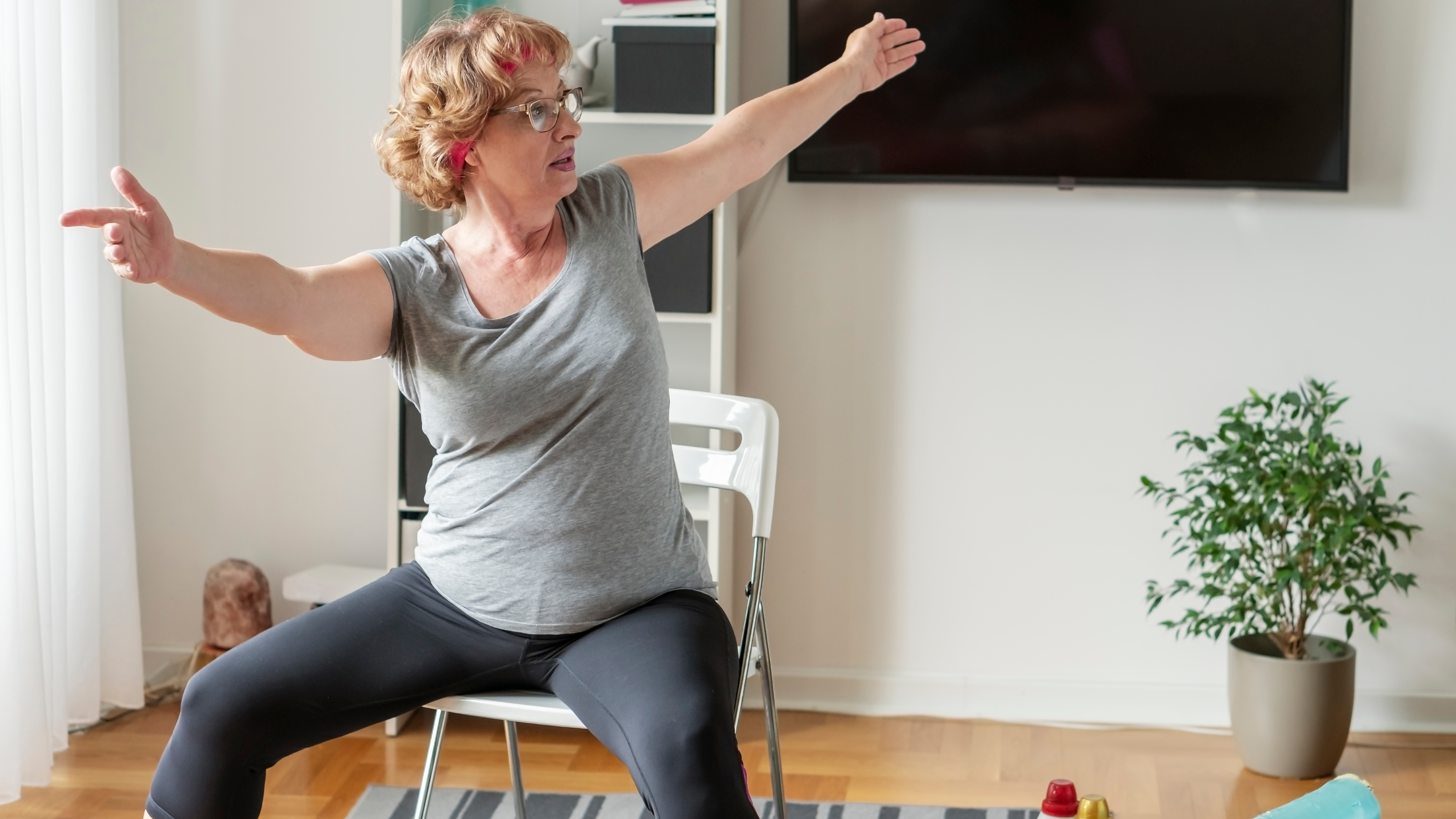 A woman sits on a plastic chair performing a stretch. Her legs are out wide, knees bent, feet on the floor, and her torso is twisting to the left as she holds her extended arms up high. Behind her we see a plant, a mounted TV and book shelves. 