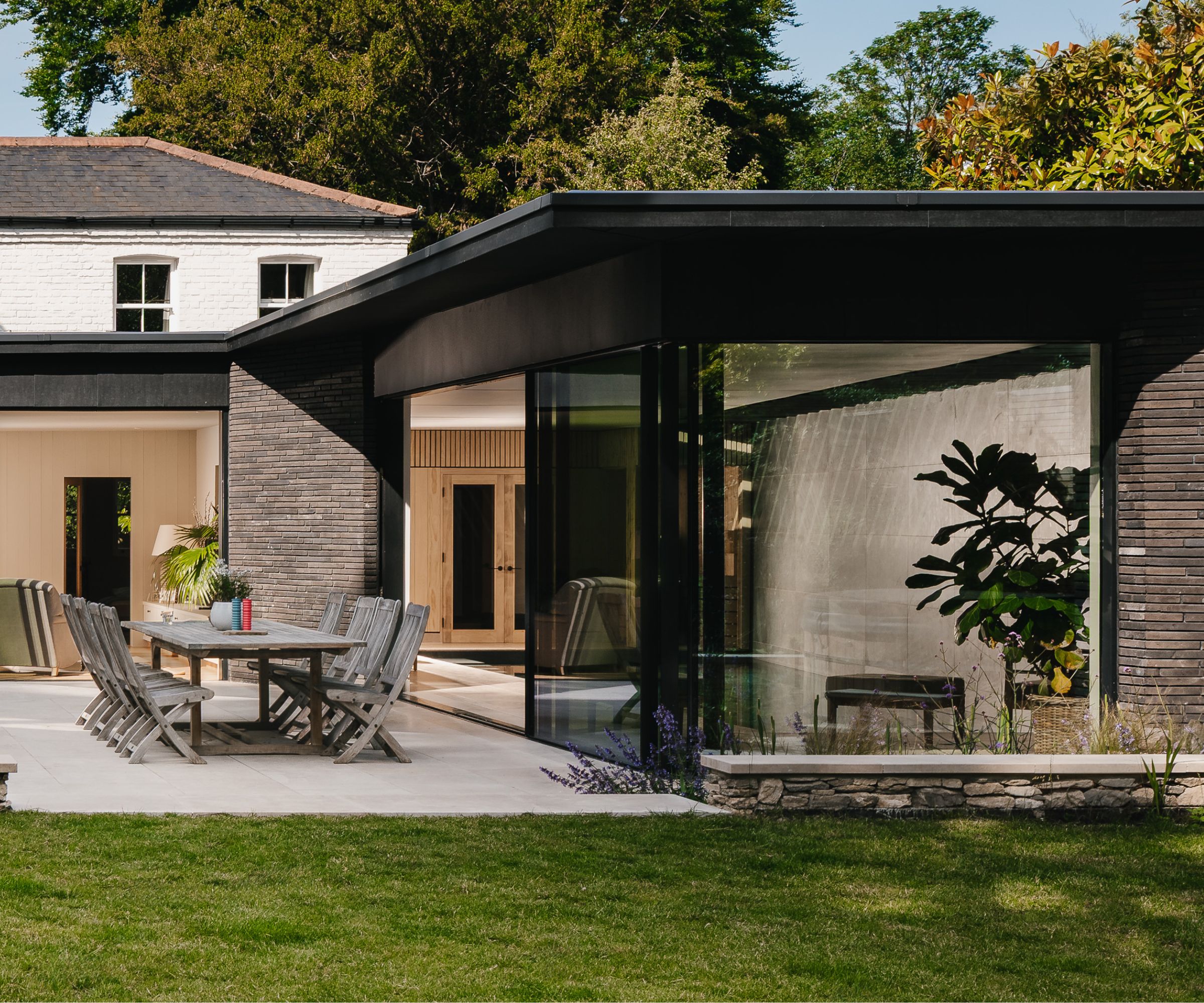 Modern extension with dark cladding, large glass panels and an outdoor dining area, blending crisp architecture with natural surroundings.