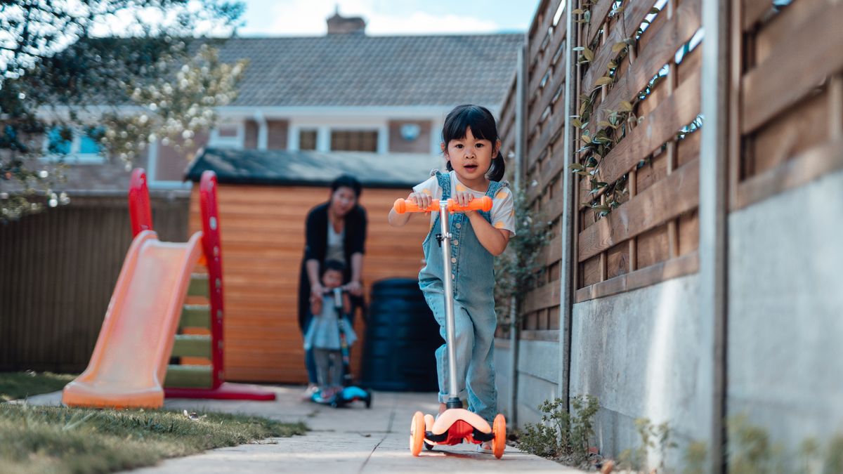 Child riding scooter in garden, with woman and another child in background