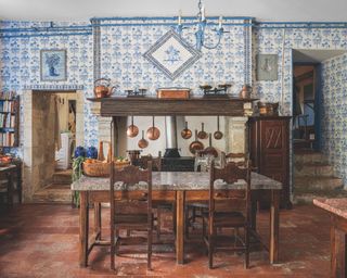 French kitchen with authentic blue and white tiled wall, fireplace, vintage furniture, terracotta tiles, copper pans, basket on marble topped table, steps to the right