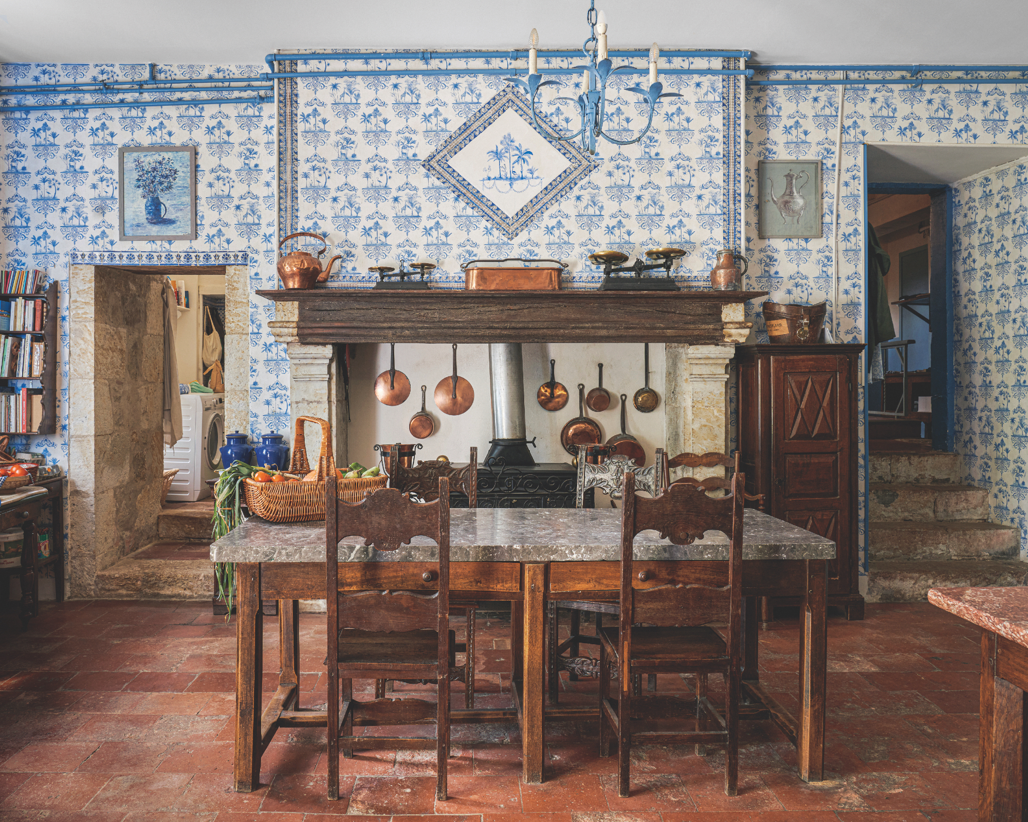 Cuisine française avec mur carrelé authentique bleu et blanc, cheminée, meubles vintage, carreaux en terre cuite, casseroles en cuivre, panier sur table en marbre, marches à droite