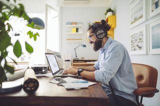 A man in a button down dress shirt works in an office setting on a laptop while wearing headphones.