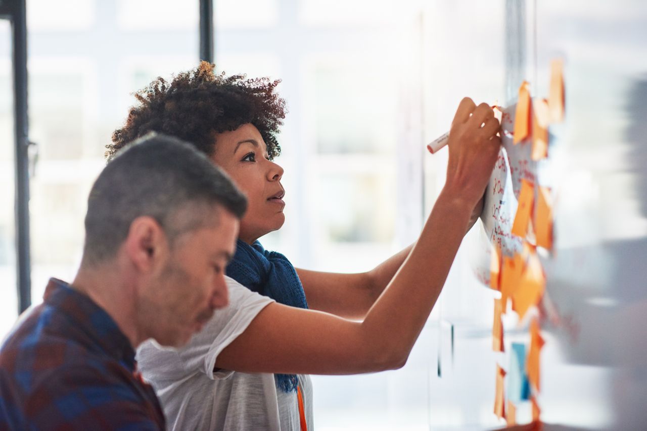 Woman and man standing in front of whiteboard covered in post-its. The woman is writing on the board with a marker.