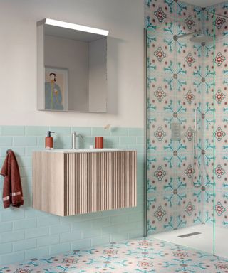 A small bathroom with a light wooden floating vanity with a silver faucet, light blue splashback tiles, a white wall with a silver cabinet above it, and a shower with pink and blue mosaic tiles