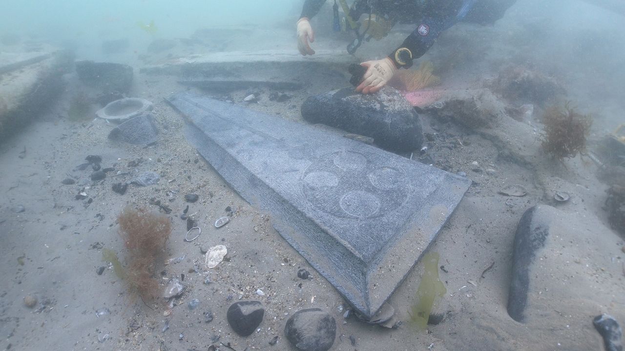 A close up a scuba diver by a gravestone with Christian designs found on the seafloor.