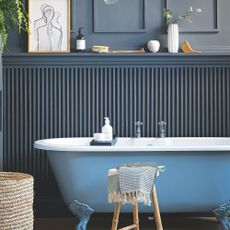 Blue and white freestanding bathtub, in front of dark blue clad wall, and a wooden stool with tassled blanket on it