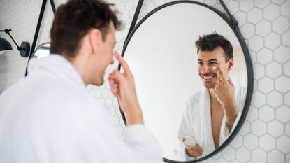 White man looking in the mirror applying a white moisturiser cream under his eyes