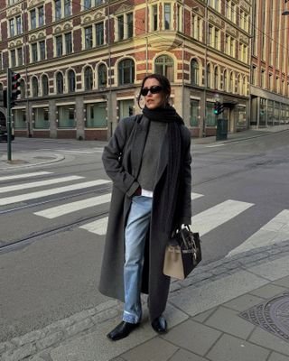 Annabel wears a gray long coat, black scarf, gray sweater, blue jeans, and black boots.