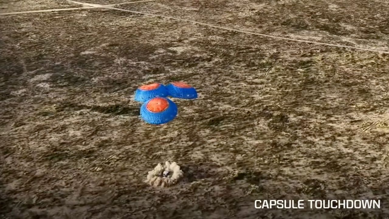 A white Blue Origin space capsule lands under blue and red parachutes.
