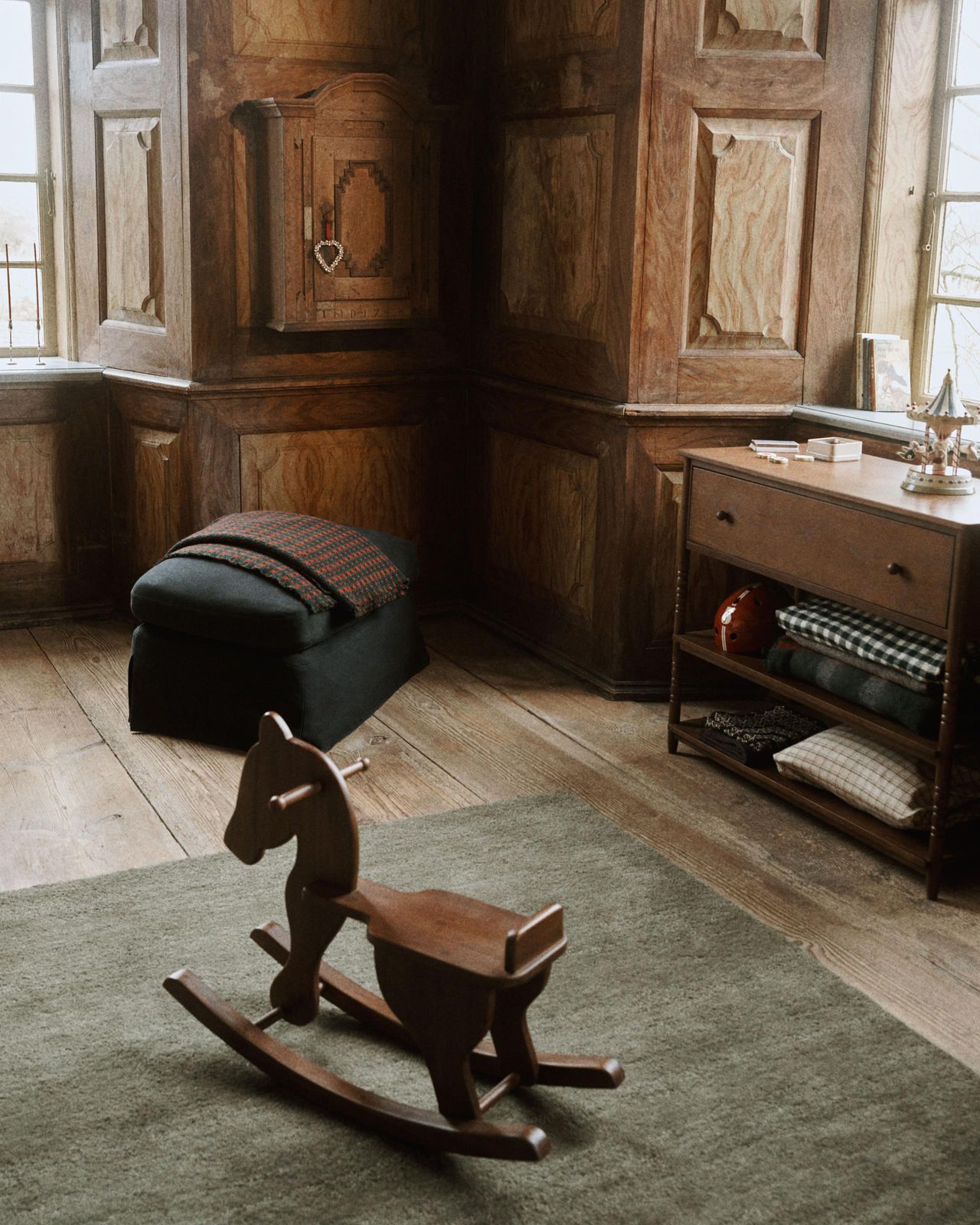 Image of a wooden room with a dark green rug that has a wooden rocking chair on it. There is a cabinet that was multiple blankets stored in it.