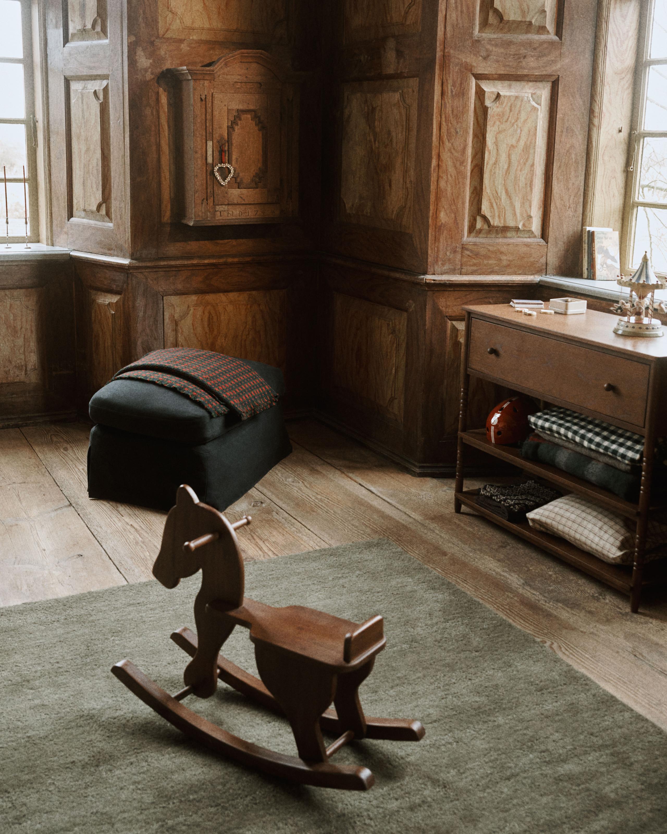Image of a wooden room with a dark green rug that has a wooden rocking chair on it. There is a cabinet that was multiple blankets stored in it.