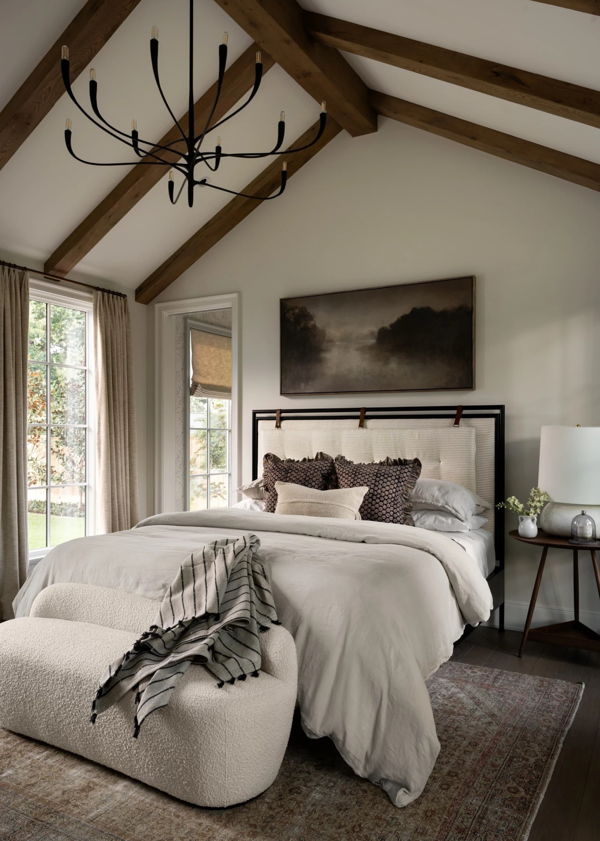 A white bedroom with wood beams on the vaulted ceiling, a black wire chandelier. a white bed with a black metal frame, layered patterned pillows, a boucle bench, a wood side table, a large area rug, and a framed painting