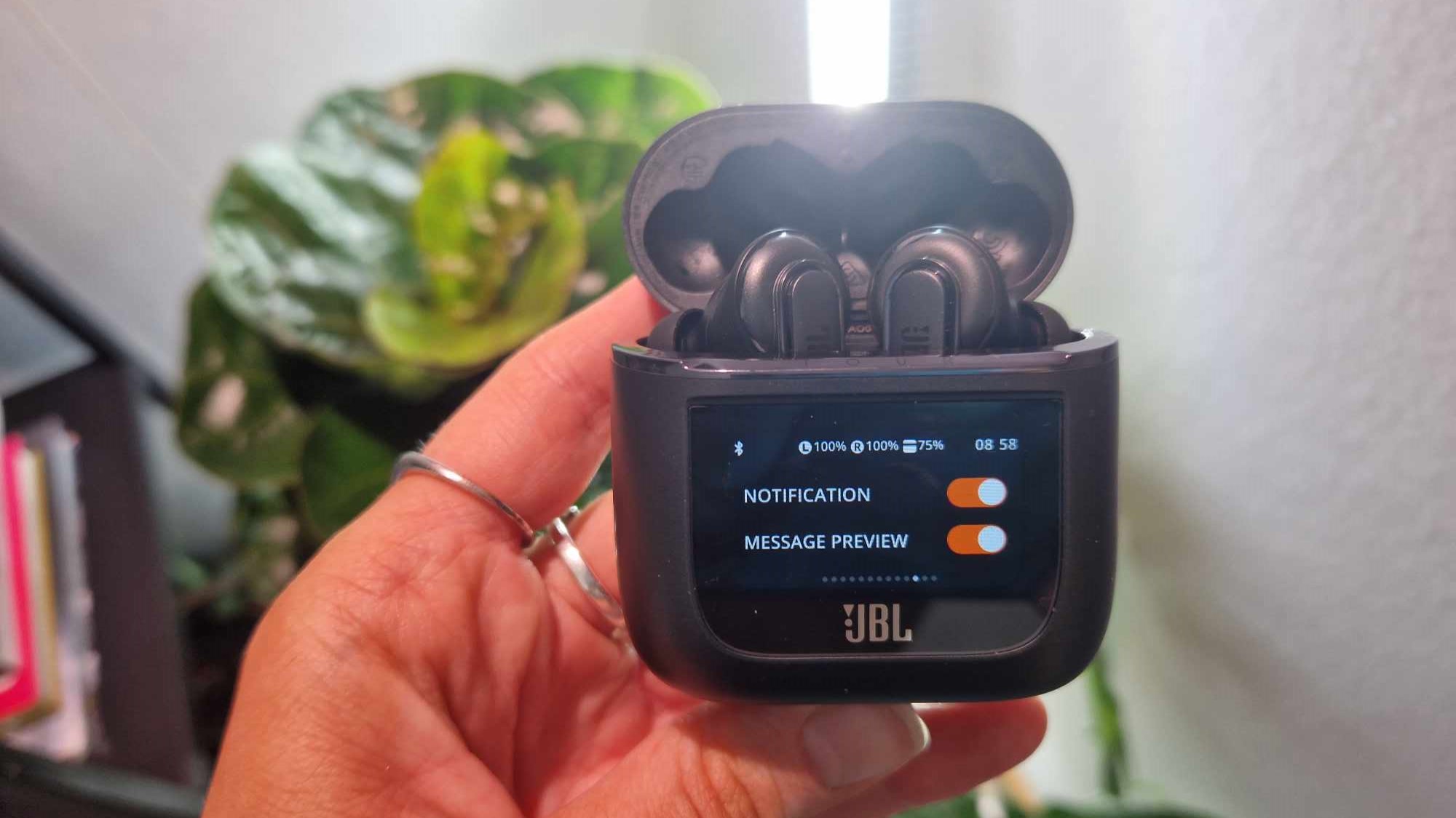 En hand håller upp ett par JBL Tour Pro 2 som ligger i sitt tillhörande laddningsfodral.