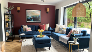 Laura's living room with a burgandy feature wall, blue sofas, wooden flooring, and lamp in the right corner