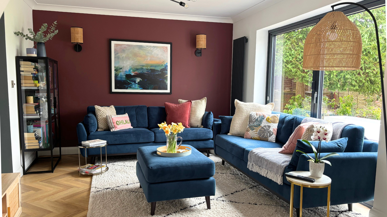 Laura's living room with a burgandy feature wall, blue sofas, wooden flooring, and lamp in the right corner