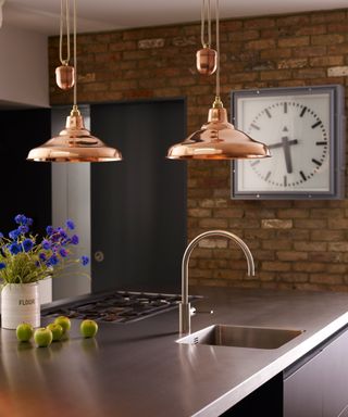 Copper rise and fall pendants above kitchen counter, with exposed brick wall and graphic wall clock in background.