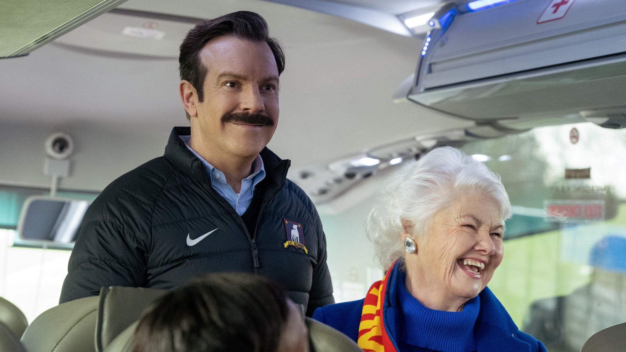 Jason Sudeikis as Ted Lasso with Annette Badland as Mae standing together on the team bus in Ted Lasso Season 4.
