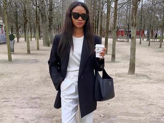 Influencer Lorna Humphrey of Symphony of Silk poses in a wooded park wearing black oversize sunglasses, a black blazer, white t-shirt, white trousers, black Prada bag, and gray New Balance sneakers.