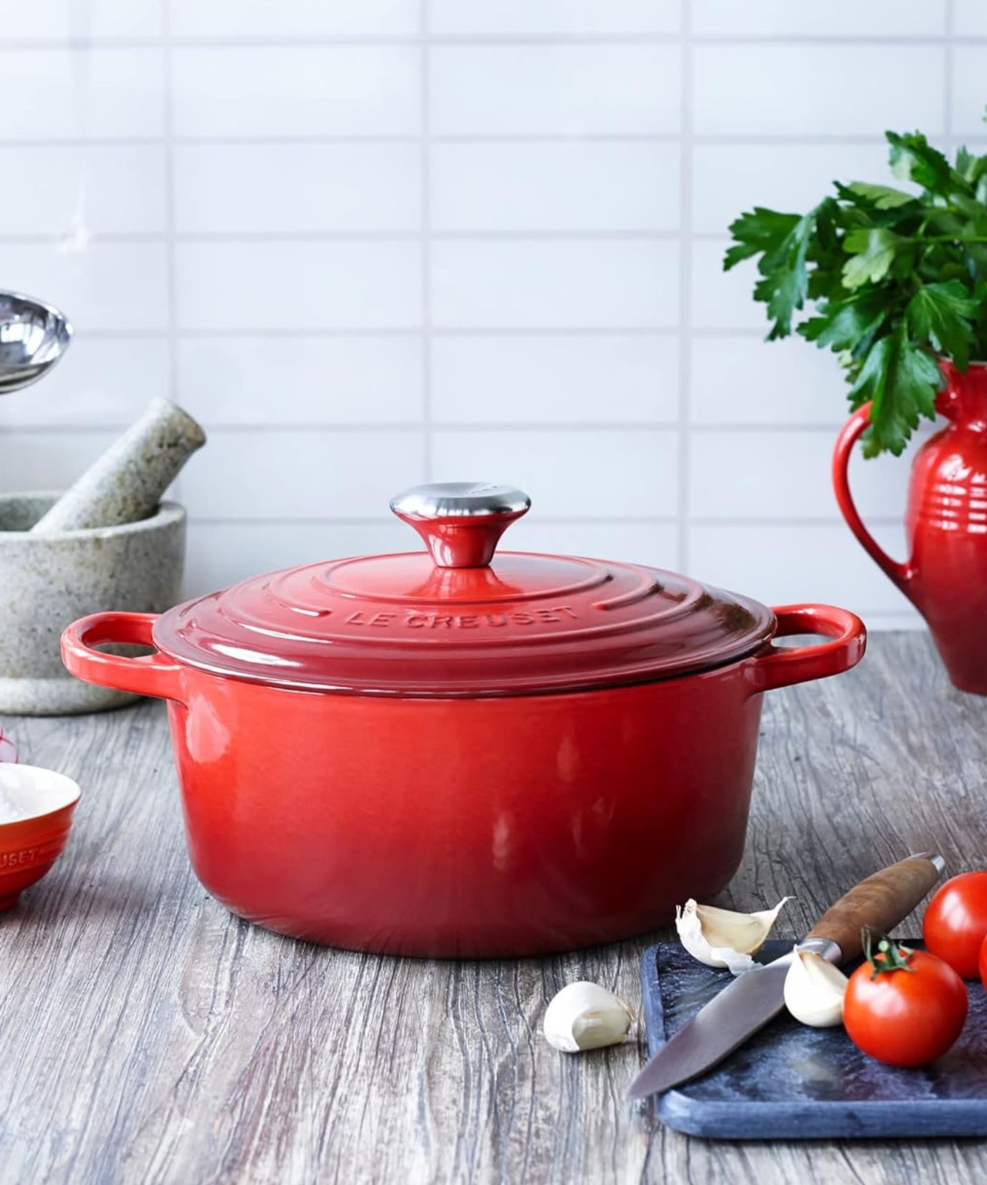 Le Creuset Enameled Cast Iron Signature Round Dutch Oven in Cerise on a gray wooden kitchen counter. There is a gray pestle and mortar, red jug with parsley, and a stone cutting board with tomatoes and garlic around the surface.