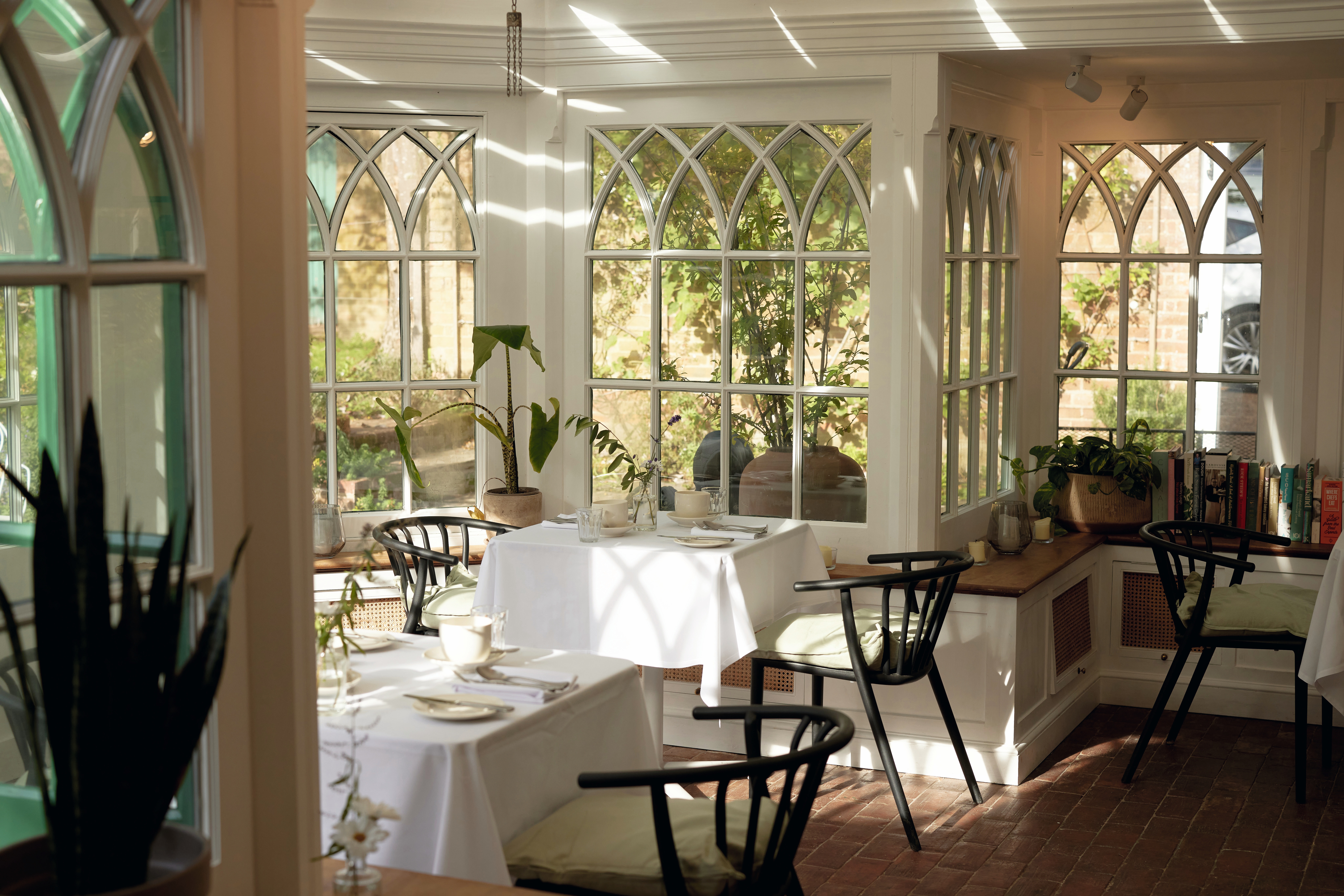 A sun-lit, bucolic restaurant dining room with Venetian-style windows, white table clothes-topped tables, and mid-century modern wooden chairs overlooks a leafy garden.
