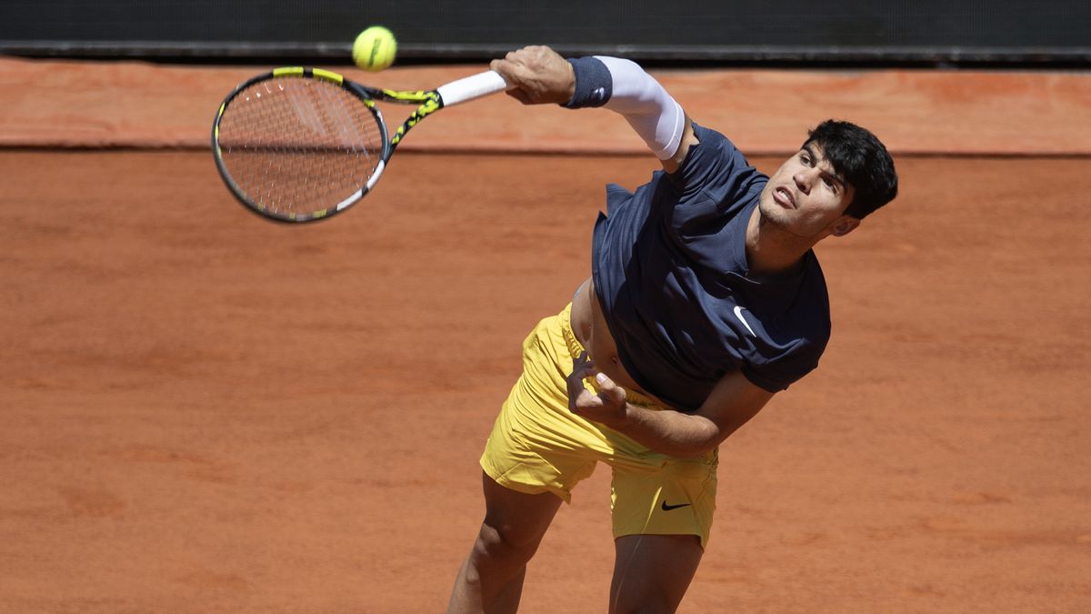 Carlos Alcaraz serves at the French Open 2024 in a Nike blue t-shirt and yellow shorts on the red clay of the Roland-Garros tennis courts