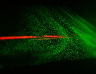 A laser beam (red) and the cloud of generated particles (illuminated by an auxiliary green laser, which makes each particle shine) in a cloud chamber.