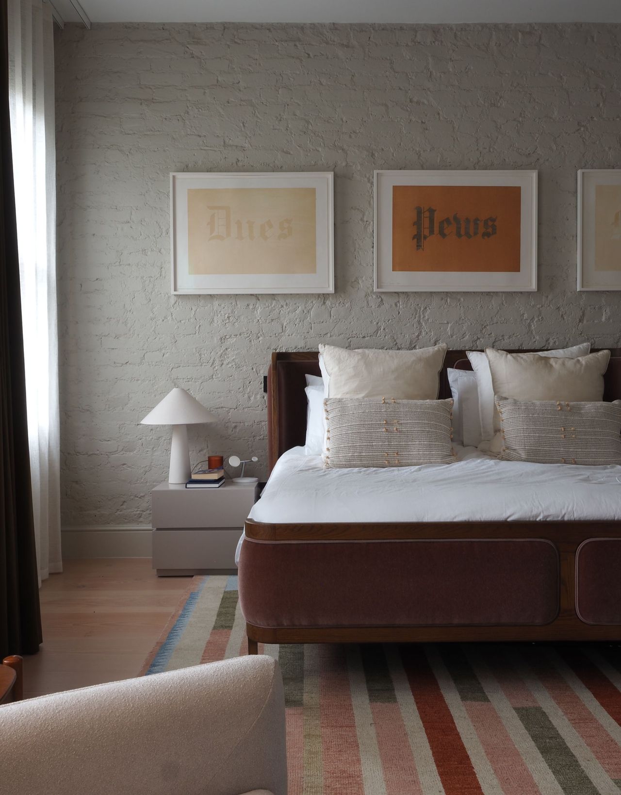 A modern bedroom with beige-painted brick walls, framed artwork above the bed and a striped patterned rug underneath the bed