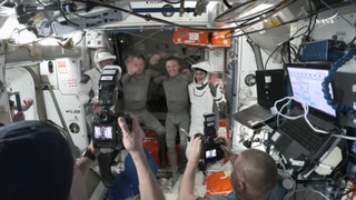 Four astronauts, three men and one woman, wave a farewell as they prepare to leave the space station. Two are in white spacesuits.