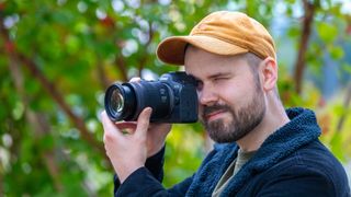 A man in an orange cap holds a Canon EOS R6 Mark II to eye level with a Canon RF 28-70mm F2.8 IS STM lens attached