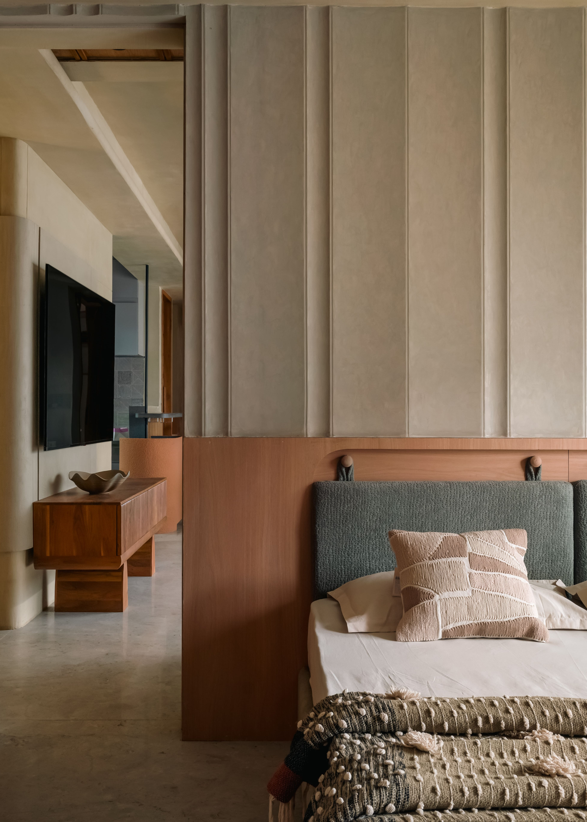 A bedroom with a cushioned headboard, patterned pillows, and a textured green throw by a door leading to a TV room