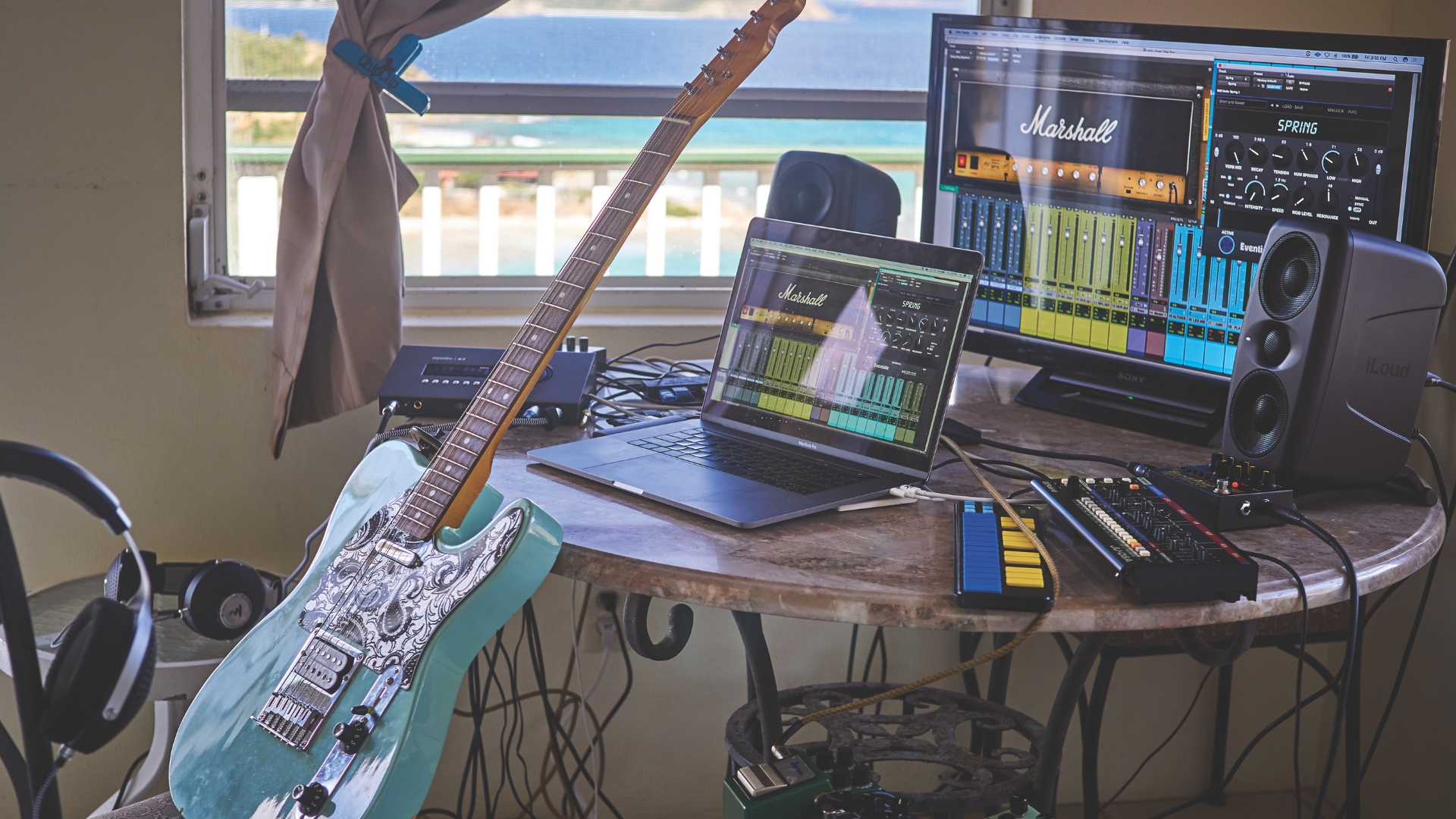 A messy studio set up with laptop, studio monitors, and electric guitar
