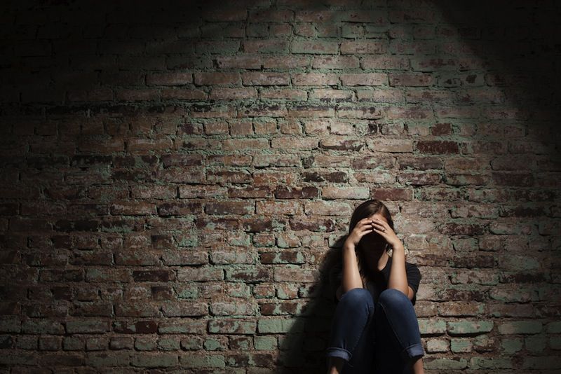 A woman sits alone near a brick wall, her face hidden in shadows.