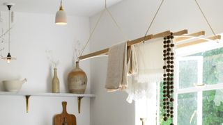 A traditional wooden ceiling-hung pulley airer suspending linen cloths and a lace-trimmed fabric in a bright kitchen with stone vases, brass shelf brackets and pendant lights.