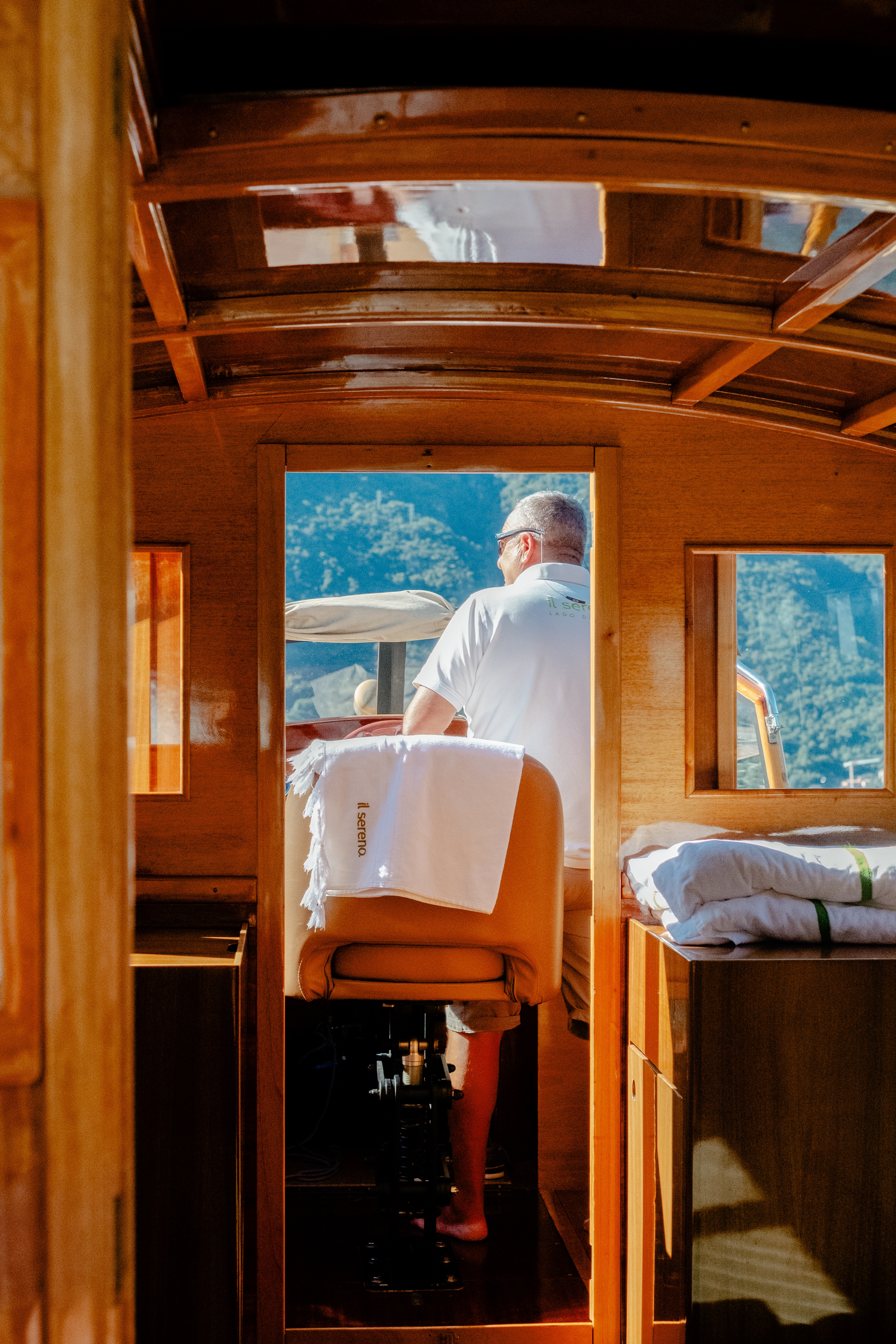 A man in a white polo shirt is photographed as he drives a sleek, lacquered wooden boat in the sunshine.