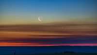 the moon shines next to bright venus at sunset. the sky is painted hues of blue, yellow and orange.