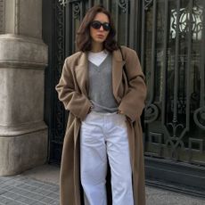 Woman wears camel coat, grey v-neck jumper, white T-shirt, white jeans and black loafers