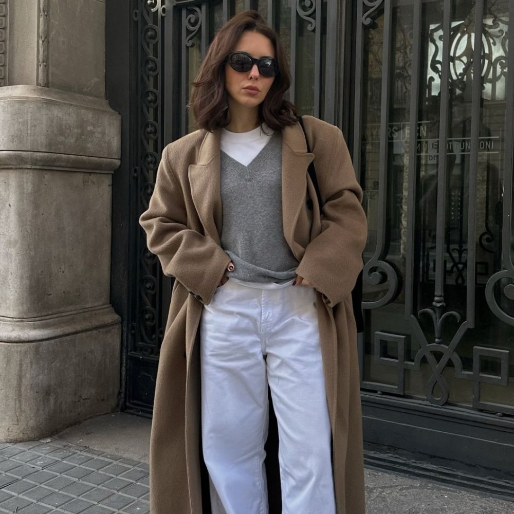 Woman wears camel coat, grey v-neck jumper, white T-shirt, white jeans and black loafers