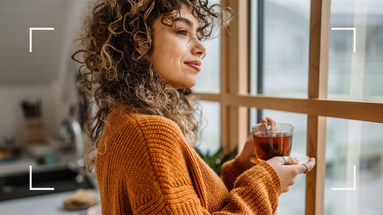 Woman looking out of the window, smiling, holding cup of herbal tea, representing why we keep waking up early